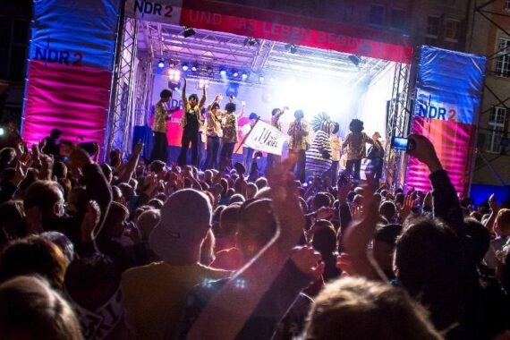 Eine lebhafte Menschenmenge erhebt ihre Hände bei einem nächtlichen Freiluftkonzert vor einer hell erleuchteten Bühne, auf der eine Band auftritt. Die Bühne ist mit Bannern mit der Aufschrift NDR 2 bedeckt.