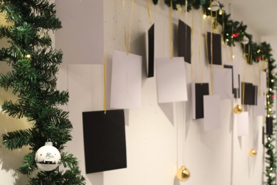 Eine Wand, die mit hängenden schwarzen und weißen Karten, goldenen Ornamenten und grüner Girlande mit Lichtern und einer weißen Christbaumkugel geschmückt ist, schafft eine festliche Urlaubsatmosphäre.