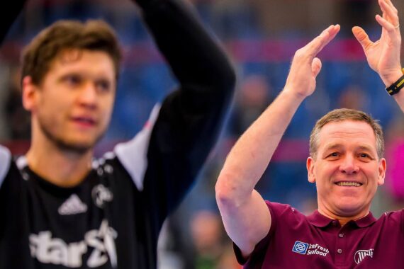 Ein lächelnder Mann in einem kastanienbraunen Hemd klatscht mit erhobenen Händen, während ein Mann in einem schwarzen Trikot neben ihm steht und ebenfalls klatscht, wobei ein unscharfer blauer Hintergrund ein Sportereignis andeutet.