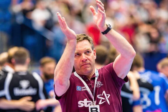 Ein Mann, der ein kastanienbraunes Polohemd mit Logos trägt, klatscht in einer Halle in die Hände, während im Hintergrund unscharfe Menschen und Spieler in Sportuniformen zu sehen sind.