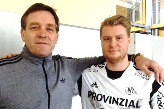 Zwei Männer stehen dicht beieinander und lächeln in die Kamera. Der Mann links trägt eine graue Jacke mit Reißverschluss, während der Mann rechts ein weißes Sporttrikot mit der Aufschrift PROVINZIAL und der Nummer 24 trägt.
