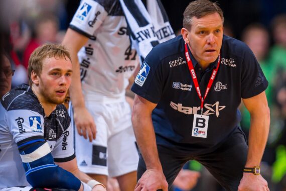 Zwei Männer in dunklen Sportuniformen, wahrscheinlich ein Trainer und ein Spieler, schauen während eines Handballspiels aufmerksam von der Seitenlinie aus zu. Der Spieler sitzt mit einem bandagierten Arm da, während der Trainer eine Trillerpfeife trägt und sich konzentriert nach vorne lehnt.