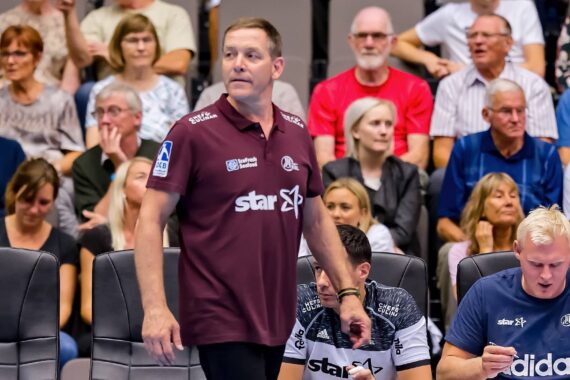 Ein Mann in einem kastanienbraunen Hemd steht bei einer Sportveranstaltung vor sitzenden Menschen, während andere Männer in Sportkleidung an einem Tisch in der Nähe sitzen. Das Publikum schaut im Hintergrund aufmerksam zu.