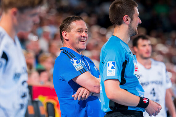 Ein lächelnder Trainer in einem blauen Hemd steht während eines Handballspiels mit verschränkten Armen neben einem Schiedsrichter, während Spieler und Zuschauer im Hintergrund verschwommen zu erkennen sind.