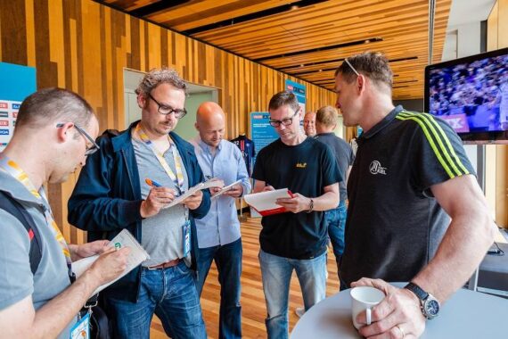 Fünf Männer stehen in einem modernen Raum zusammen, diskutieren und machen sich Notizen. Einige halten Notizblöcke und Stifte in der Hand, eine Person hält eine Kaffeetasse. Im Hintergrund ist ein Fernseher mit einem Sportereignis zu sehen.