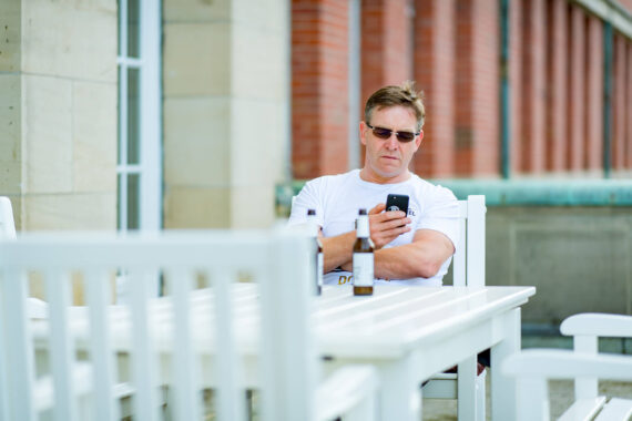 Ein Mann mit Sonnenbrille sitzt an einem weißen Tisch im Freien und schaut auf sein Telefon. Auf dem Tisch steht eine Bierflasche, und im Hintergrund ist ein Gebäude mit roten Ziegeln und Steinmauern zu sehen.