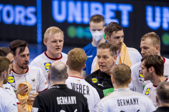 Eine Herren-Handballmannschaft in weißen Trikots, einige mit der Aufschrift GERMANY, schart sich während eines Spiels um einen Trainer. Eine Person trägt eine Maske. Im Hintergrund ist das Wort UNIBET auf einer blauen Anzeigetafel zu sehen.