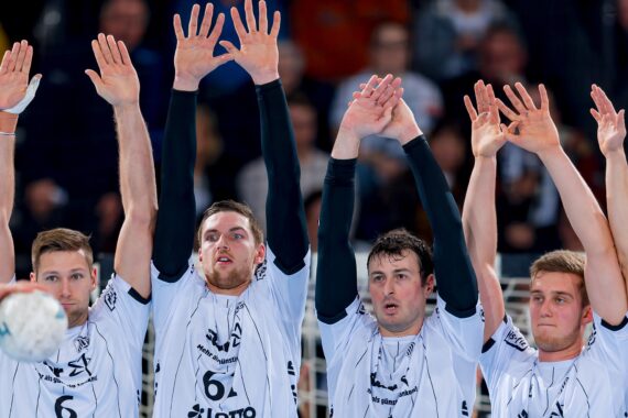 Vier männliche Handballspieler in weißen Trikots stehen mit erhobenen Armen in einer Reihe und bilden eine Abwehrmauer während eines Spiels. Ein Spieler hält einen Handball. Im Hintergrund ist eine unscharfe Menschenmenge zu sehen.