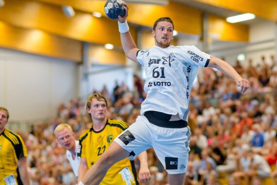 Ein Handballspieler in einem weißen Trikot springt, um den Ball zu werfen, während drei Verteidiger in Gelb ihn beobachten. Das Spiel findet in einer mit Zuschauern gefüllten Halle statt.