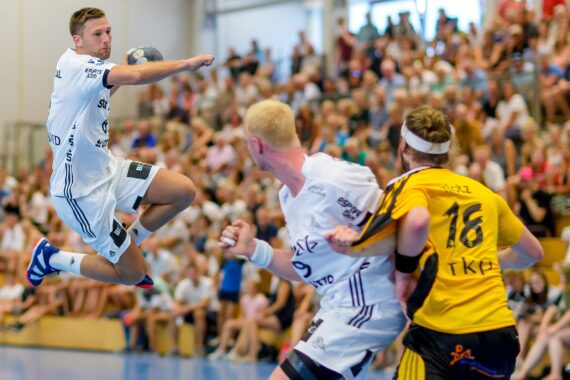 Ein Handballspieler in einem weißen Trikot springt in die Luft, um den Ball zu werfen, während zwei Spieler, einer in Weiß und einer in Gelb, den Ball verteidigen. Eine Zuschauermenge verfolgt das Spiel im Hintergrund.