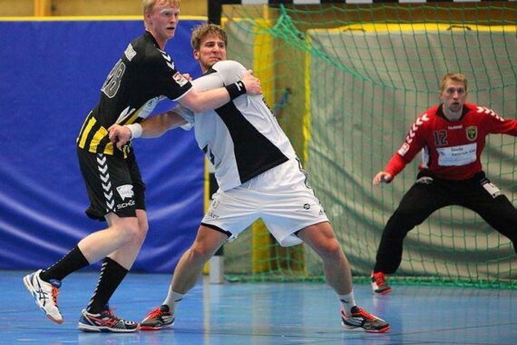 Zwei männliche Handballspieler, einer in Schwarz und einer in Weiß, kämpfen um eine Position in Tornähe, während ein Torwart in Rot vom Torraum aus aufmerksam zusieht.