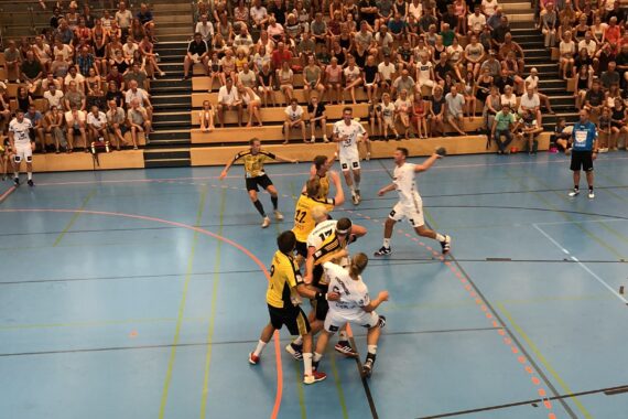 Ein Handballspiel in einer Sporthalle mit Spielern in gelben und weißen Trikots, umgeben von einem großen Publikum, das auf Tribünen sitzt. In der Nähe des Tores herrscht ein reges Treiben.
