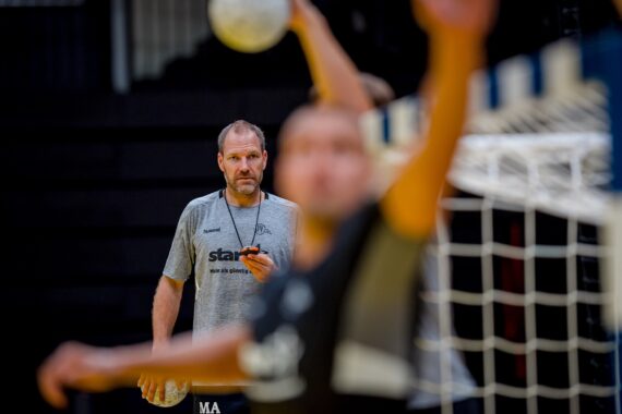 Ein Trainer, der eine Trillerpfeife in der Hand hält, beobachtet aufmerksam ein Handballtraining, mit verschwommenen Spielern und einem Tornetz im Vordergrund.