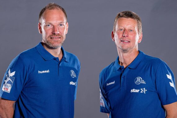 Zwei Männer in passenden blauen Sporthemden stehen nebeneinander vor einem einfarbigen grauen Hintergrund, schauen beide in die Kamera und lächeln leicht. Auf den Hemden sind verschiedene Logos zu sehen.