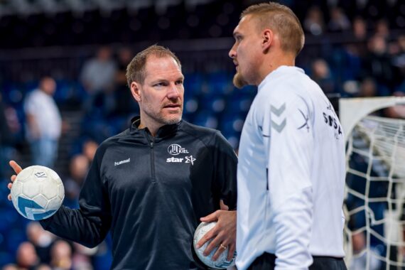 Zwei Männer in Sportkleidung stehen in einem Innenraum in der Nähe eines Torpfostens, halten Handbälle und führen ein konzentriertes Gespräch. Im Hintergrund ist ein unscharfes Publikum in einem Stadion zu sehen.