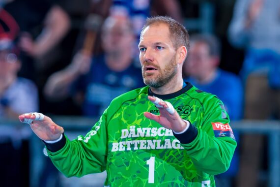 Ein männlicher Handballspieler in einem grünen Torwarttrikot mit der Aufschrift "DÄNISCHES BETTENLAGER" steht mit erhobenen Händen und wirkt während eines Spiels konzentriert. Im Hintergrund sind unscharfe Zuschauer zu sehen.