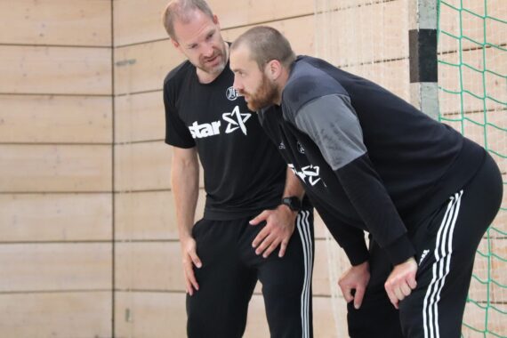 Zwei Männer in Sportkleidung stehen in einem Innenraum in der Nähe eines Netzes und führen ein ernstes Gespräch. Der eine lehnt sich nach vorne und stützt die Hände auf die Knie, während der andere mit ihm spricht und mit der Hand gestikuliert. Beide tragen schwarze Sporthemden und -hosen.