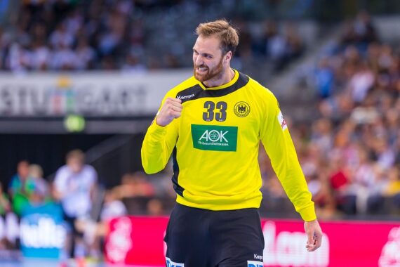 Ein männlicher Handballtorwart in einem gelben Trikot mit der Nummer 33 ballt die Faust und lächelt beim Jubeln auf dem Spielfeld, während im Hintergrund eine unscharfe Menschenmenge zu sehen ist.