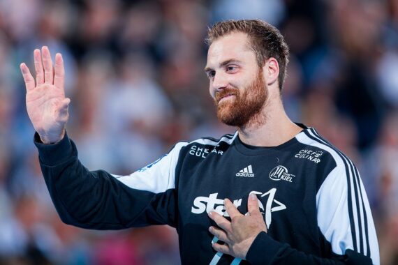 Ein bärtiger Mann in einem schwarz-weißen Sporttrikot winkt mit der rechten Hand und stützt seine linke Hand lächelnd auf seine Brust, während im Hintergrund eine verschwommene Menschenmenge zu sehen ist.