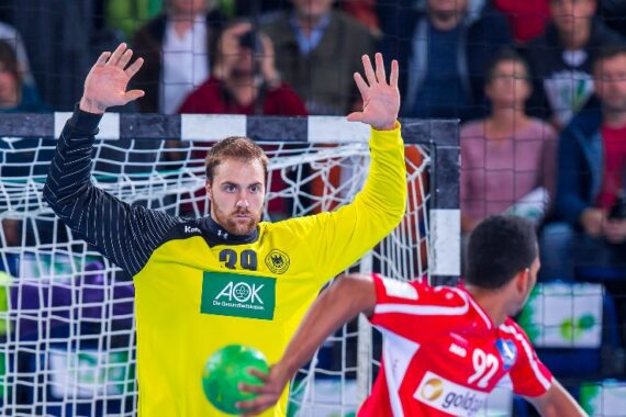 Ein Handballtorhüter in einem leuchtend gelben Trikot steht mit erhobenen Armen bereit, das Tor zu verteidigen, während ein gegnerischer Spieler in einem roten Trikot sich darauf vorbereitet, den Ball zu werfen. Die Zuschauer schauen von den Tribünen im Hintergrund zu.