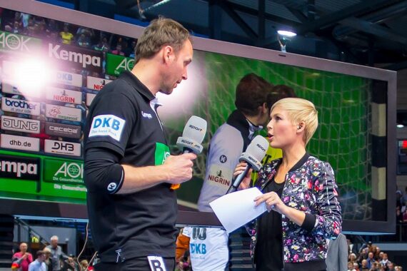 Ein Mann in Sportkleidung wird von einer Frau interviewt, die ein Mikrofon und Papiere in der Hand hält, während im Hintergrund eine große Leinwand und Werbetafeln bei einer Sportveranstaltung zu sehen sind.