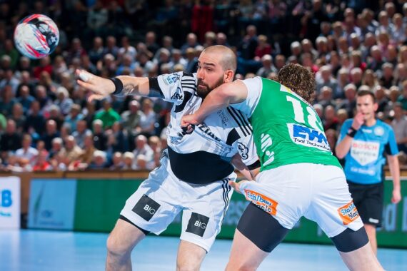 Ein Handballspieler in einem weiß-schwarzen Trikot wirft den Ball, während er von einem Gegner in einem grün-weißen Trikot während eines Spiels eng verteidigt wird, mit einer Menge von Zuschauern im Hintergrund.