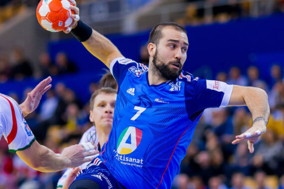 Ein männlicher Handballspieler in einem blauen Trikot mit der Nummer 7 bereitet sich darauf vor, den Ball während eines Spiels zu werfen. Er ist in Bewegung, während seine Gegner in weißen Trikots versuchen, ihn zu blockieren. Im Hintergrund sind unscharfe Zuschauer zu sehen.