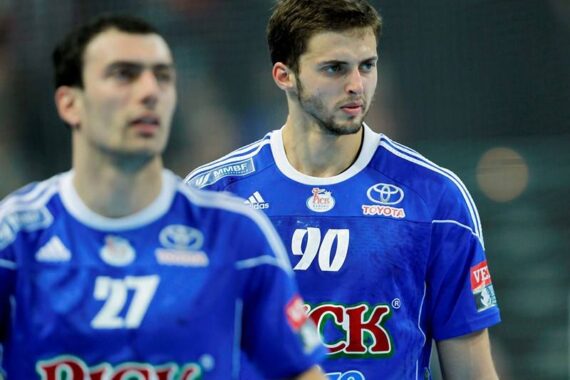 Zwei männliche Handballspieler in blauen Trikots, einer mit der Nummer 90 im Fokus, der andere mit der Nummer 27 im Vordergrund, während eines Spiels. Auf den Trikots sind mehrere Sponsorenlogos zu sehen.