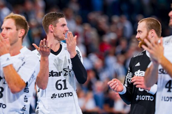 Vier Handballspieler in weißen und schwarzen Trikots stehen nebeneinander, klatschen und lächeln, mit einer unscharfen Menge im Hintergrund. Die sichtbaren Trikotnummern sind 13, 61 und 4.