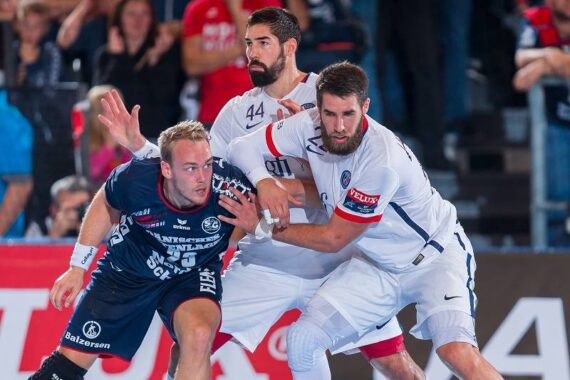 Drei männliche Handballspieler in Aktion während eines Spiels, wobei ein Spieler in einem dunkelblauen Trikot den Ball hält und zwei Spieler in weißen Trikots ihn eng verteidigen. Im Hintergrund sind Zuschauer zu sehen.