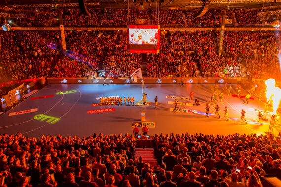 Eine große Halle, gefüllt mit jubelnden Fans, die ein hell erleuchtetes Handballspiel verfolgen, mit Mannschaften und Maskottchen auf dem Spielfeld und Pyrotechnik, die die Szene beleuchtet.