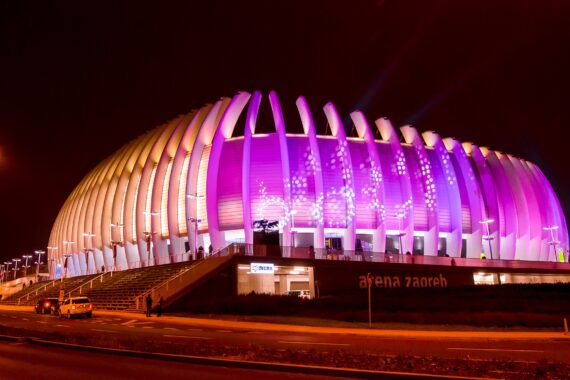 Die Arena Zagreb, ein modernes Stadion, wird nachts mit leuchtenden violetten und rosa Lichtern angestrahlt, die das charakteristische geschwungene architektonische Design und die vielen beleuchteten Säulen zur Geltung bringen. Entlang des Vordereingangs sind Autos geparkt.