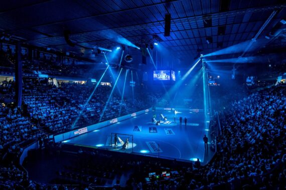 Eine überfüllte Sporthalle mit einem blau beleuchteten Spielfeld und dramatischen Scheinwerfern, gefüllt mit Zuschauern, die ein laufendes Spiel verfolgen. Große Bildschirme zeigen Live-Bilder über dem Spielfeld.