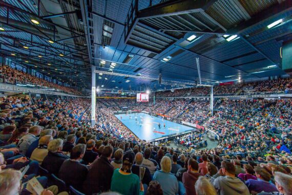Eine große Menschenmenge füllt eine Halle und verfolgt ein Handballspiel auf einem hell erleuchteten Platz. Die Tribünen sind voll mit Zuschauern, und die Decke ist mit freiliegenden Balken und Beleuchtung versehen. Über dem Spielfeld hängt eine Anzeigetafel.