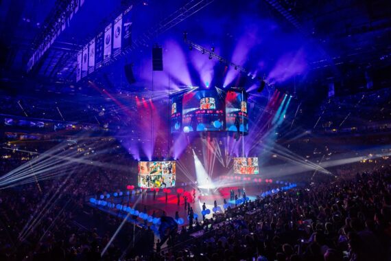 Eine lebhafte Arena mit einem jubelnden Publikum, beleuchtet von bunten Bühnenscheinwerfern, großen Videoleinwänden und Bannern über dem Kopf während eines Live-Auftritts oder einer Großveranstaltung.
