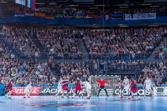 Eine überfüllte Halle mit Zuschauern, die ein Handballspiel verfolgen; Spieler in weißen und roten Trikots sind in der Nähe des Tores in Aktion, während mehrere internationale Flaggen über den Zuschauern hängen.