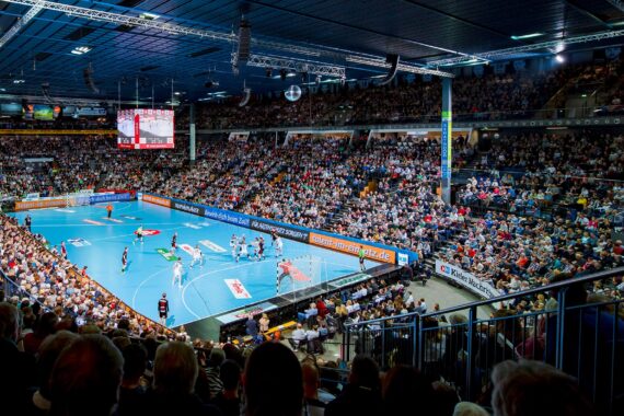 Eine große, mit Zuschauern gefüllte Halle, in der ein Handballspiel auf einem blauen Spielfeld stattfindet, wobei die Spieler in der Nähe eines der Tore aktiv sind. Die Lichter des Stadions erhellen die Szene hell.