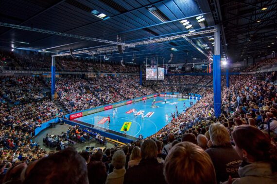 Eine große, überfüllte Halle mit Zuschauern, die ein Handballspiel zwischen zwei Mannschaften auf einem blauen Spielfeld verfolgen. Helle Lichter beleuchten die Spielfläche, und große Bildschirme zeigen das Geschehen über dem Spielfeld.