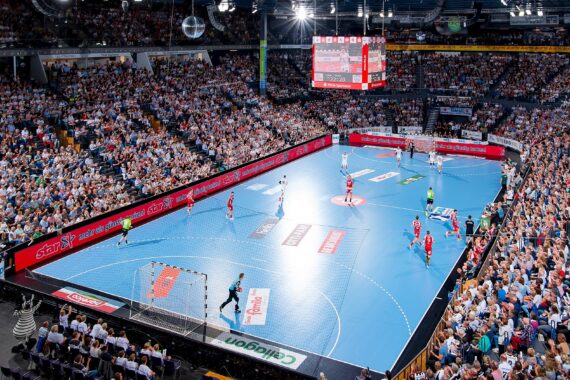 Eine große Halle, gefüllt mit Zuschauern, die ein professionelles Handballspiel auf einem blauen Spielfeld verfolgen, mit Spielern in roten und weißen Trikots und einer Anzeigetafel über der Mitte.