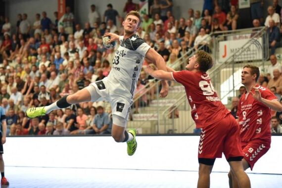 Ein Handballspieler in einem weißen Trikot springt in der Luft, um den Ball zu werfen, während zwei Spieler in Rot versuchen, ihn zu blockieren. Die Szene spielt sich in einer überfüllten Halle ab.