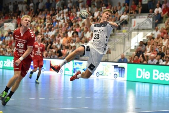 Ein männlicher Handballspieler in einem weißen Trikot springt in die Luft, um den Ball zu werfen, während ein Spieler in einem roten Trikot hinter ihm herläuft. Ein großes Publikum verfolgt das Spiel in einer Halle.