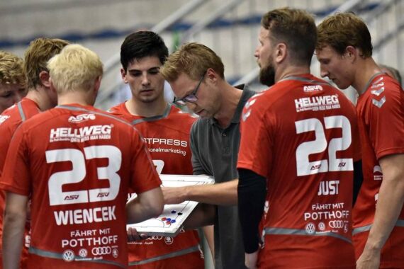 Ein Trainer mit Brille bespricht während einer Auszeit die Strategie mit einer Gruppe männlicher Handballspieler in roten Trikots, von denen einige die Nummer 23 tragen. Die Spieler hören aufmerksam zu, während der Trainer auf ein Whiteboard zeigt.