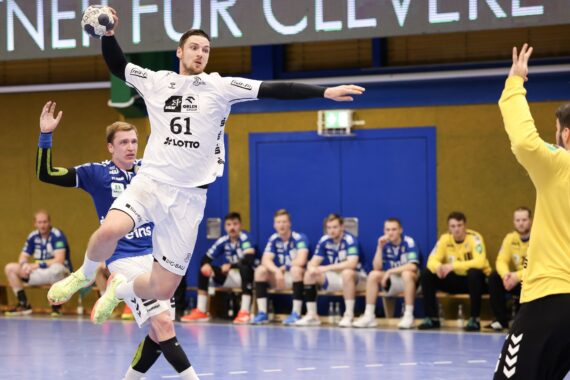 Ein Handballspieler in einem weißen Trikot springt in der Luft, um den Ball auf das Tor zu werfen, während die Gegner in Blau den Ball verteidigen und der Torwart in Gelb sich darauf vorbereitet, den Schuss abzublocken; die Mannschaftskameraden schauen von der Bank aus zu.