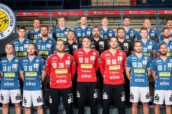 Eine Männer-Handballmannschaft stellt sich in der Halle in drei Reihen auf. Sie trägt blau-weiße Trikots, bis auf vier Spieler in Rot. In der oberen linken Ecke ist ein kreisförmiges Logo des Erzgebirgischen Handballvereins Aue zu sehen.