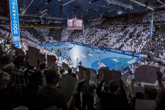 Eine überfüllte Halle mit Zuschauern, die weiße Zettel hochhalten und ein Handballspiel auf einem blauen Spielfeld unter grellem Licht verfolgen. Die Atmosphäre ist lebhaft und energiegeladen.