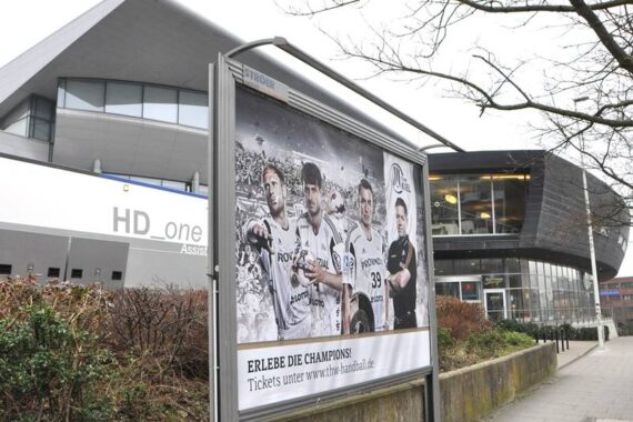 Eine Plakatwand zeigt fünf Männer in Sportuniformen mit dem Text ERLEBE DIE CHAMPIONS! vor einem modernen Gebäude mit großen Fenstern und dem Zeichen HD_one. Der Himmel ist bedeckt und die Bäume sind blattlos.
