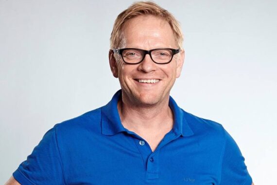 Ein lächelnder Mann mittleren Alters mit blondem Haar und schwarzer Brille trägt ein hellblaues Poloshirt. Er steht vor einem schlichten hellen Hintergrund.