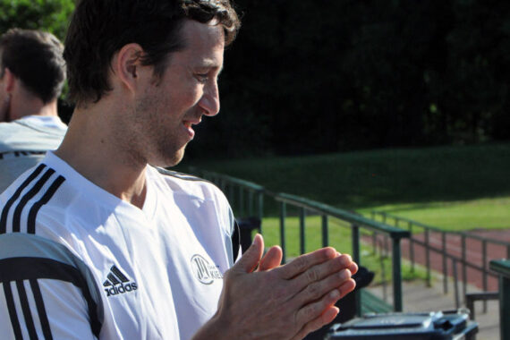 Ein Mann in einem weißen Adidas-Sporthemd steht im Freien, lächelt und klatscht in die Hände, während im Hintergrund eine Laufbahn und grünes Gras zu sehen sind.