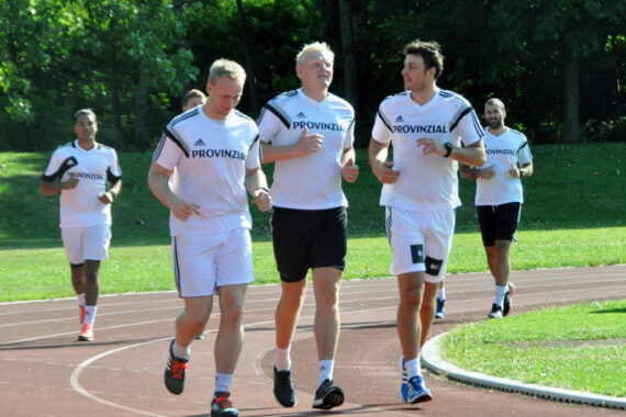 Eine Gruppe von Männern in passenden weißen Sportuniformen joggt gemeinsam auf einer Bahn, umgeben von Grün und Bäumen im Hintergrund.