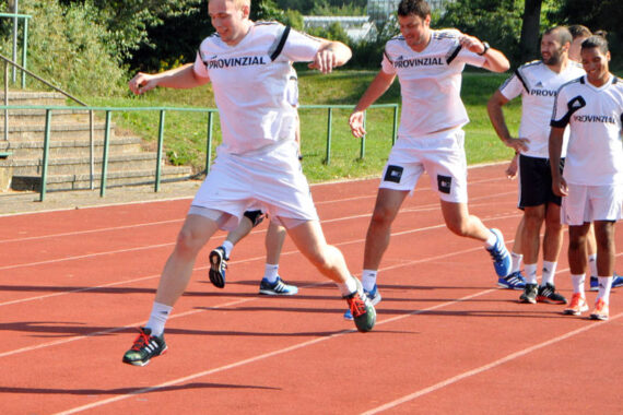 Mehrere Männer in weißen Sportuniformen springen auf einer roten Laufbahn im Freien, mit Grünflächen und Stufen im Hintergrund. Ein Mann ist mitten im Sprung, während andere zusehen oder sich auf den Sprung vorbereiten.